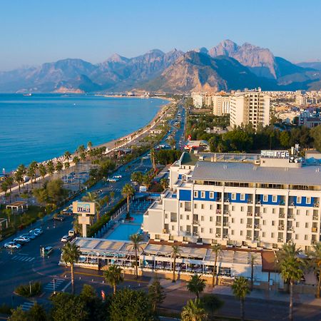 אנטליה Sealife Family Resort Hotel מראה חיצוני תמונה The photo depicts a scenic coastal view, featuring a beachfront area lined with palm trees. In the foreground, there is a hotel or resort with a pool and outdoor seating. The road runs parallel to the beach, with some vehicles visible. In the backgro