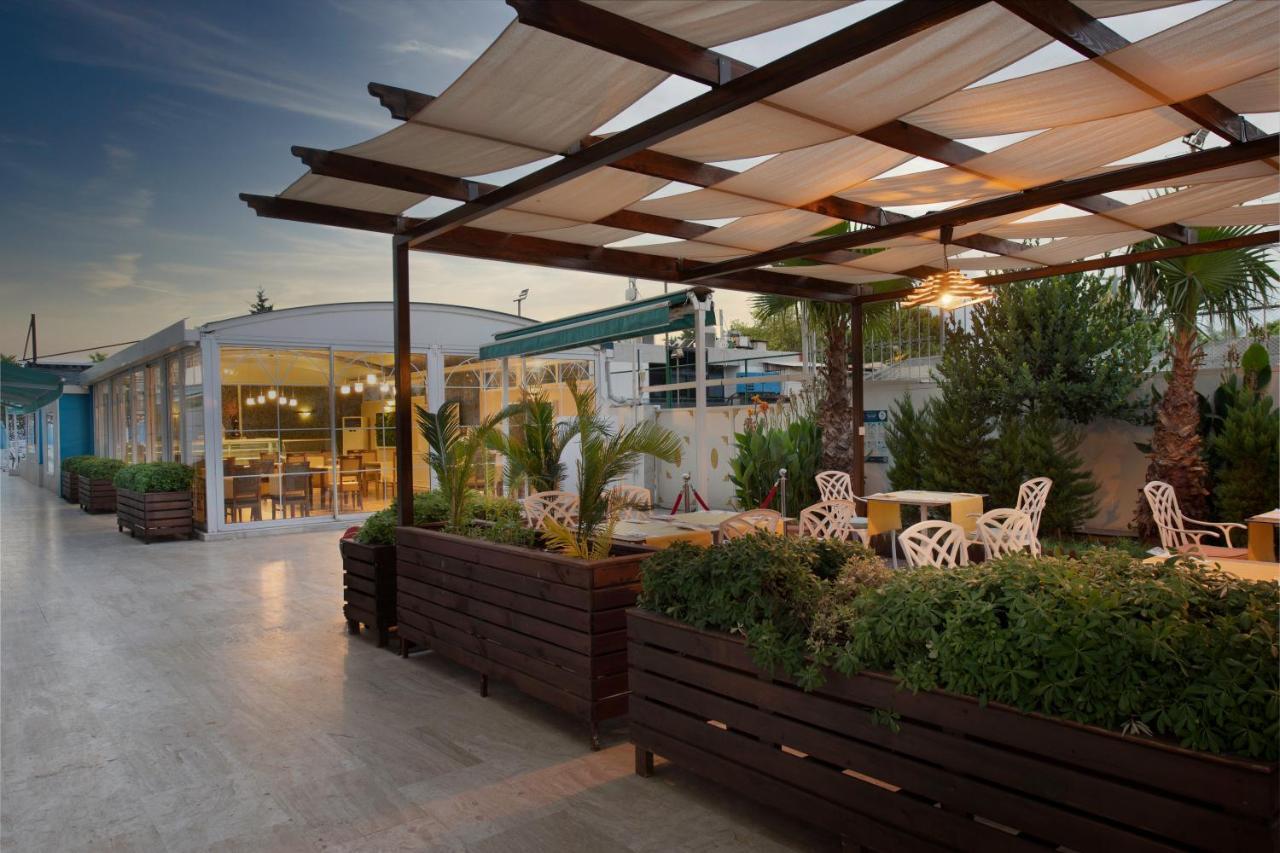אנטליה Sealife Family Resort Hotel מראה חיצוני תמונה The photo depicts an outdoor space, likely a café or restaurant terrace. It features a wooden structure with a grid-like canopy that provides shade, and several white chairs with decorative designs are arranged around small tables. There are large pl