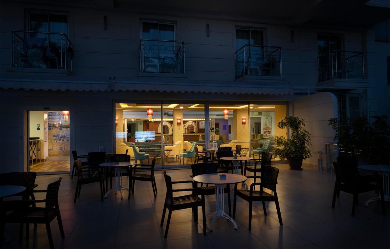 אנטליה Sealife Family Resort Hotel מראה חיצוני תמונה The photo depicts an outdoor seating area of a café or restaurant. In the foreground, there are several black chairs and a small round table. In the background, through large windows, you can see the interior of the establishment, which is warmly lit