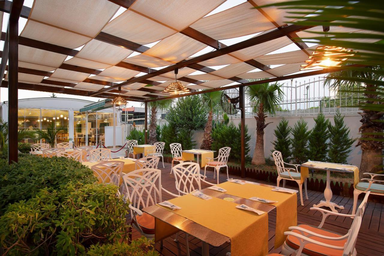אנטליה Sealife Family Resort Hotel מראה חיצוני תמונה The photo depicts an outdoor dining area of a restaurant featuring a pergola with fabric coverings that provide shade. The space is arranged with several tables adorned with bright yellow tablecloths. The chairs are designed with a distinctive white 