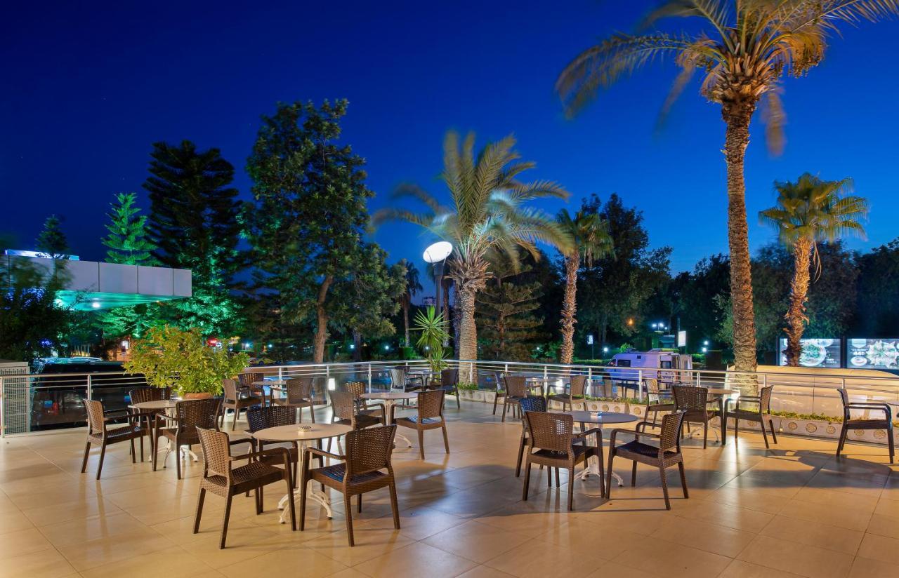 אנטליה Sealife Family Resort Hotel מראה חיצוני תמונה The photo depicts an outdoor dining area, likely part of a restaurant or café, set in the evening. There are several tables with chairs arranged across a tiled patio. The space is surrounded by tall palm trees and greenery, creating a tropical ambian