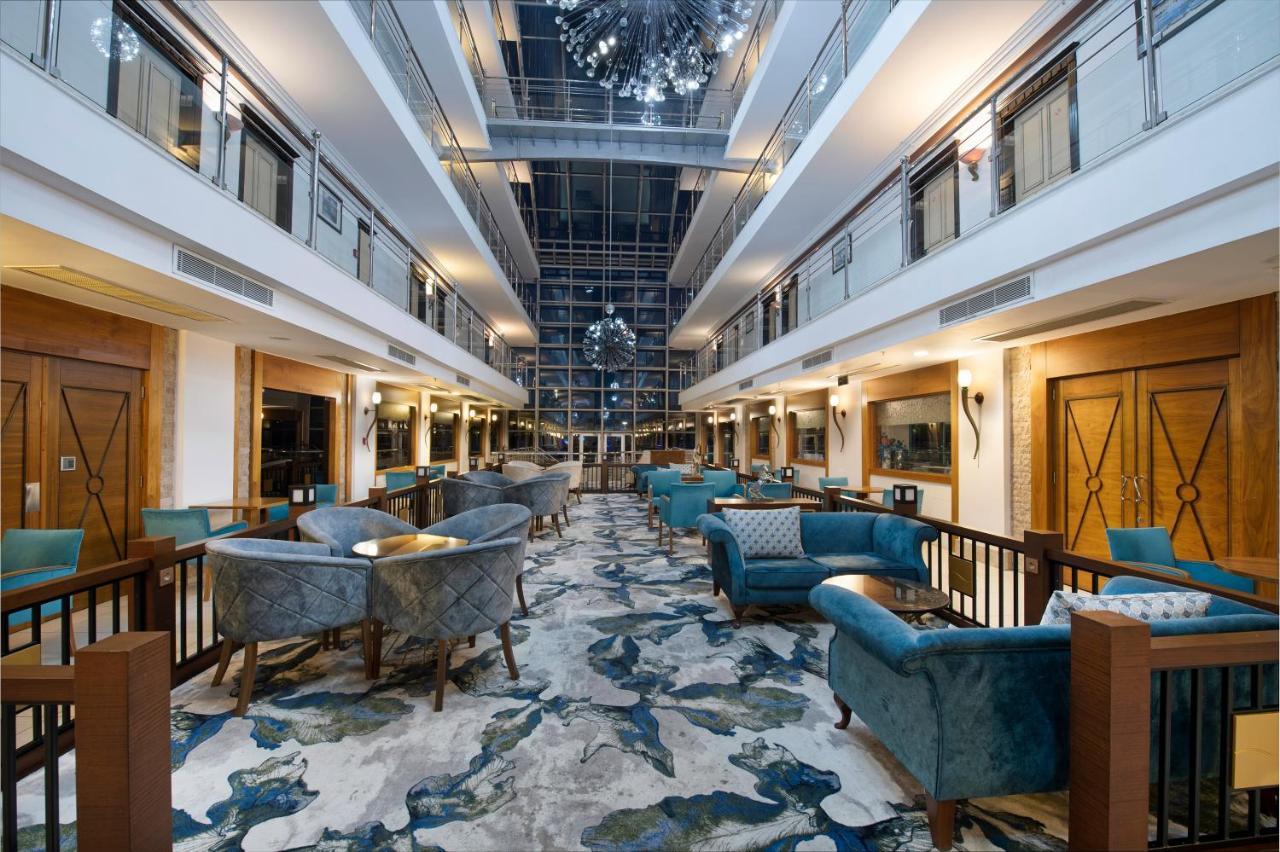 אנטליה Sealife Family Resort Hotel מראה חיצוני תמונה The photo shows a spacious and elegantly designed hotel lobby. There are two levels with balconies overlooking the main area. The lobby features plush, stylish seating arranged around small tables. The floor is decorated with a colorful, abstract car