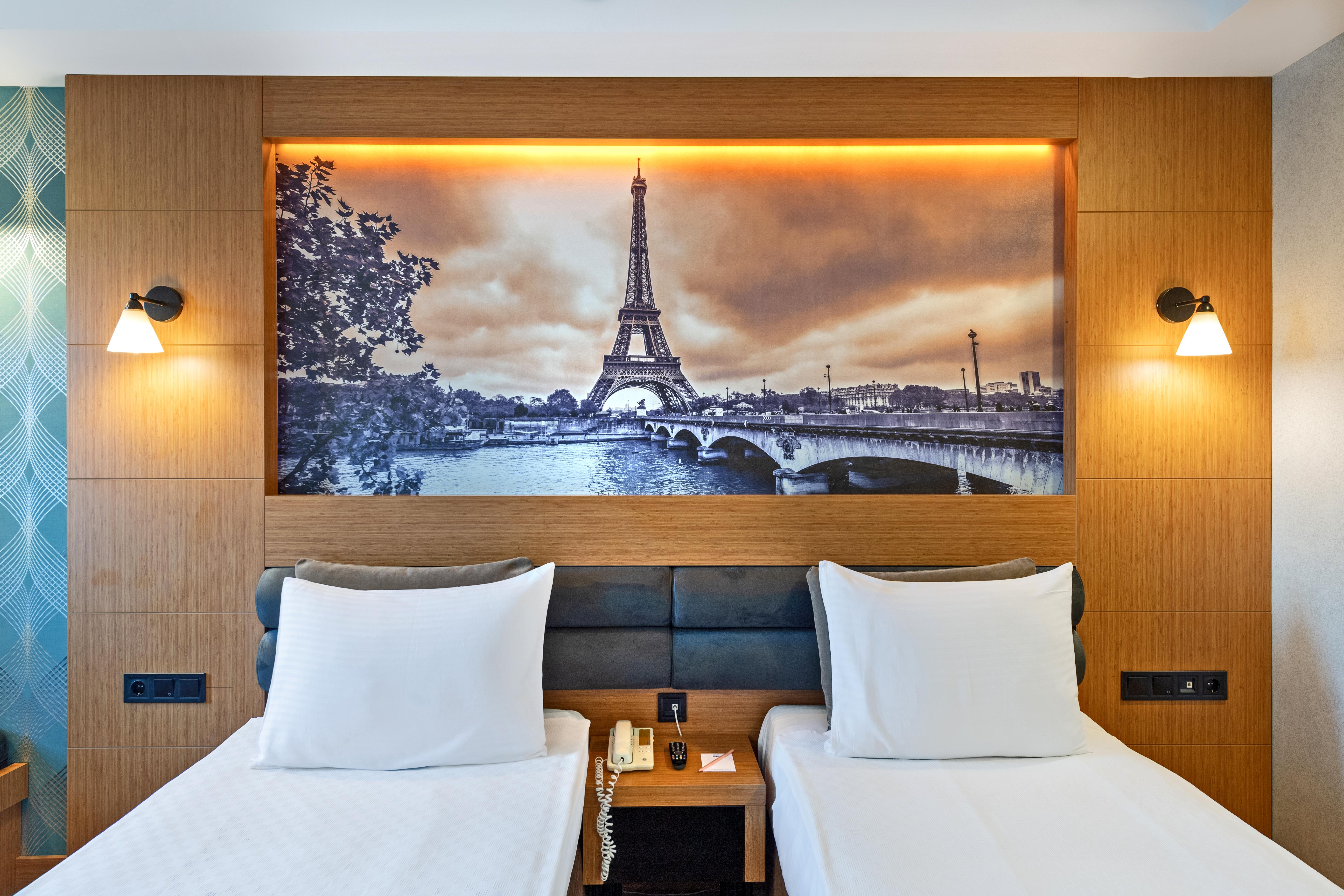 אנטליה Sealife Family Resort Hotel מראה חיצוני תמונה The photo depicts a hotel room featuring two single beds with white bedding. Behind the beds, there's a large mural of the Eiffel Tower, set against a scenic backdrop of the Seine River. The image has a vintage, sepia-toned appearance, enhancing the 