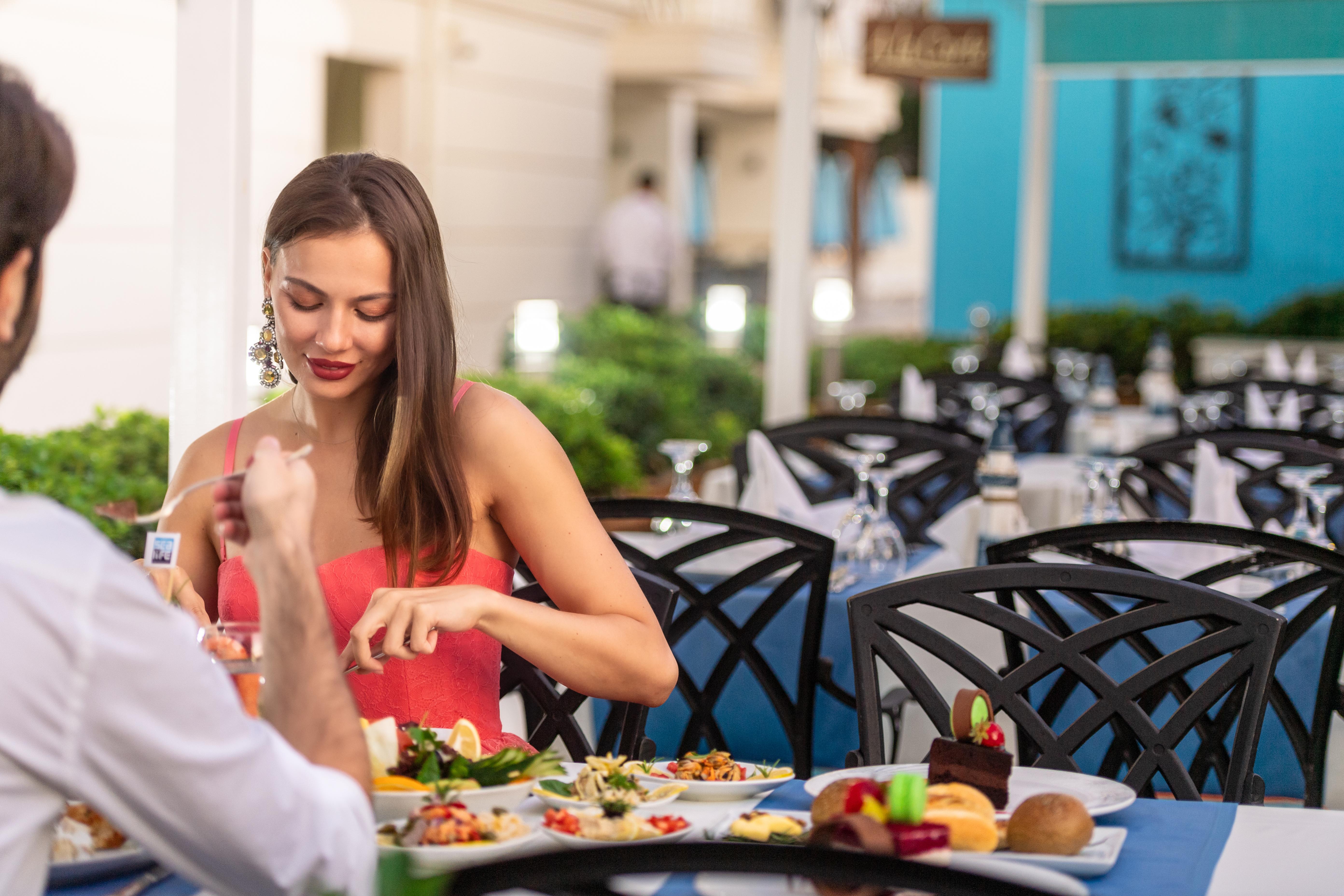 אנטליה Sealife Family Resort Hotel מראה חיצוני תמונה The photo shows a dining scene at a restaurant. A woman in a red dress is seated at a table, interacting with her meal. She appears to be taking time to enjoy her food. The table is set with various dishes, including salads and desserts. The backgrou