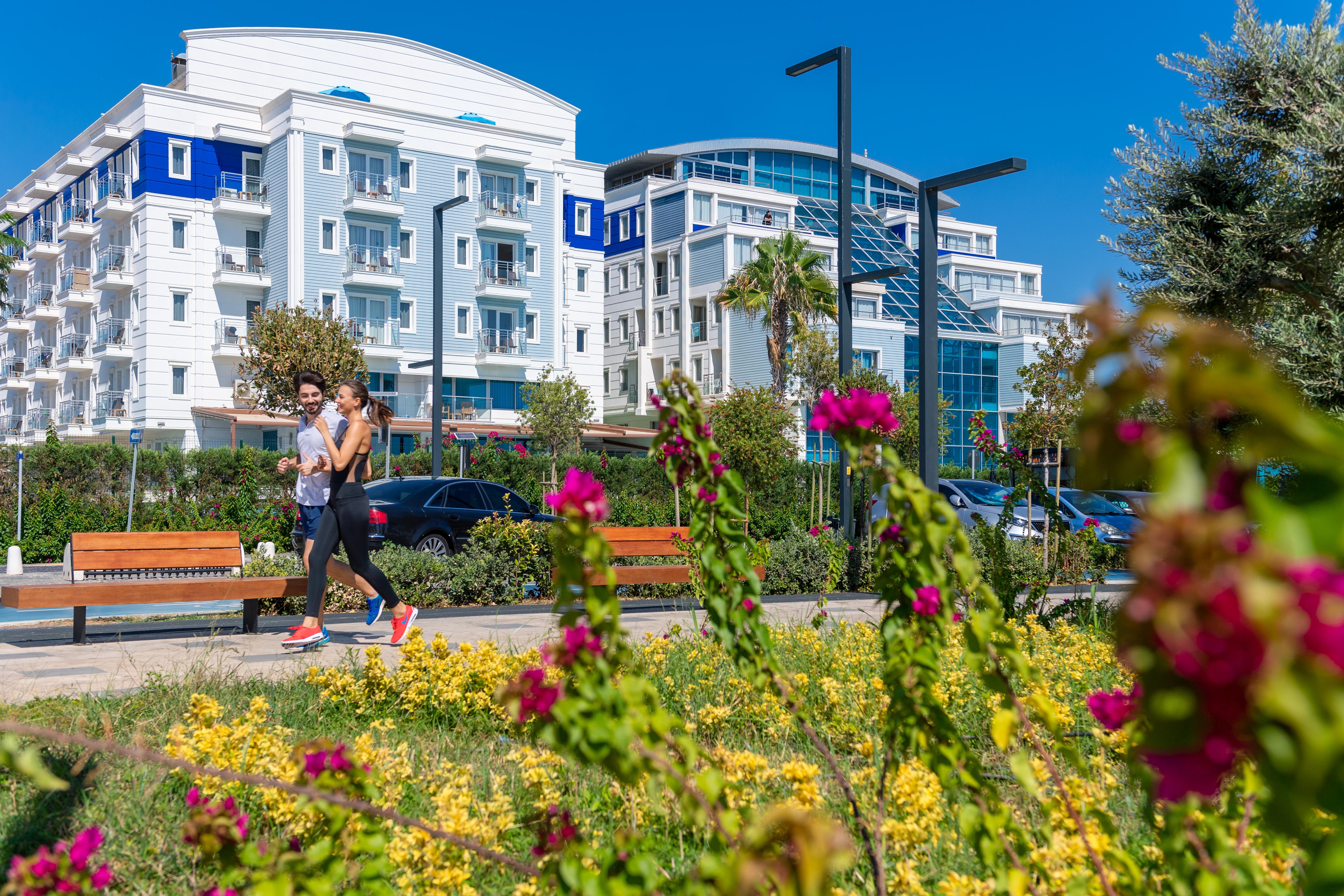 אנטליה Sealife Family Resort Hotel מראה חיצוני תמונה The photo depicts a sunny outdoor scene featuring two individuals walking along a path. They are surrounded by a vibrant landscape filled with yellow flowers and pink bougainvillea. In the background, there are modern buildings with balconies, some h