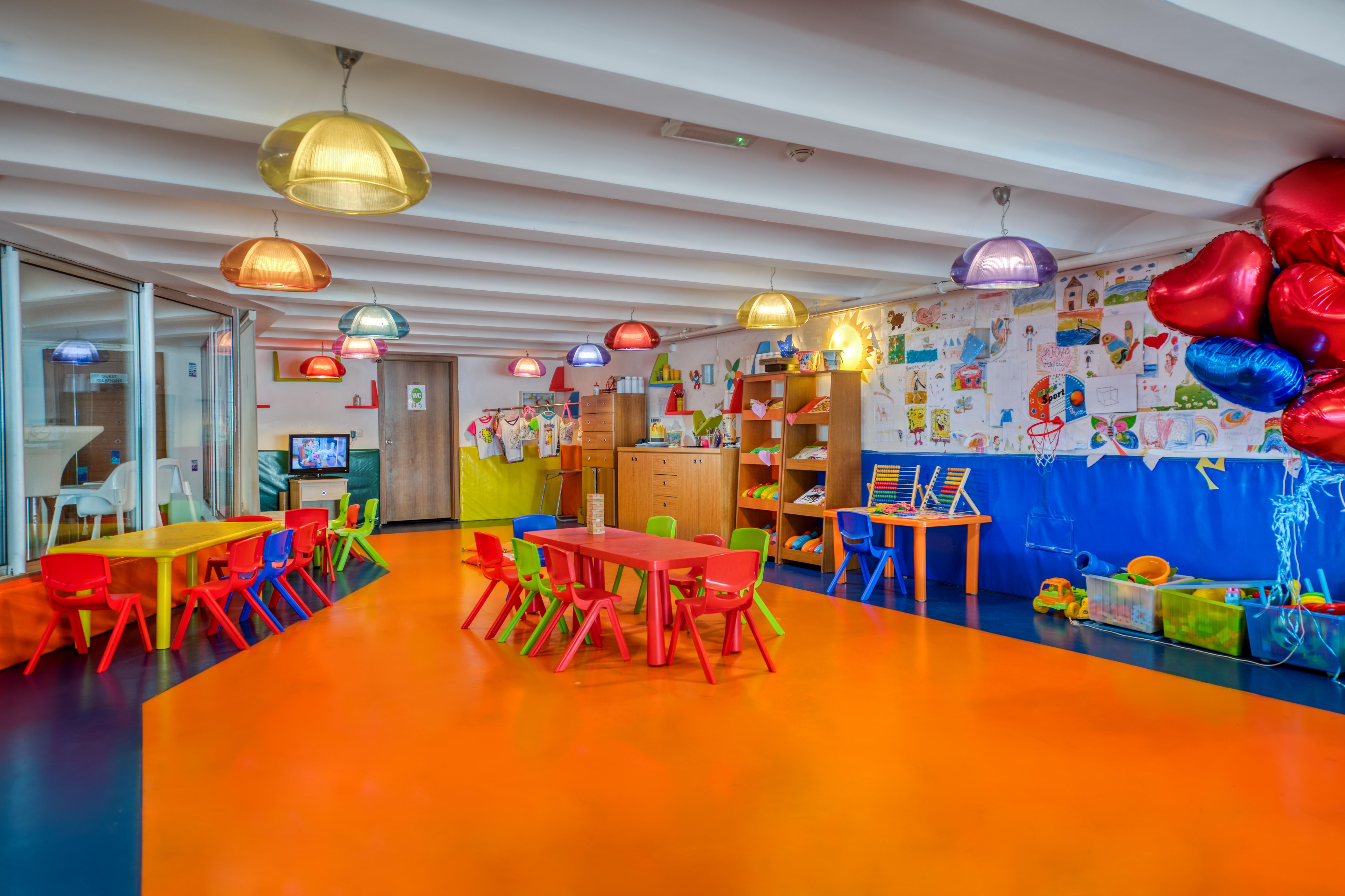 אנטליה Sealife Family Resort Hotel מראה חיצוני תמונה The photo shows a vibrant and colorful kindergarten classroom. The flooring is a bright orange, contributing to the cheerful atmosphere. Various tables and chairs are arranged throughout the space, with some in yellow and red. Colorful pendant lights