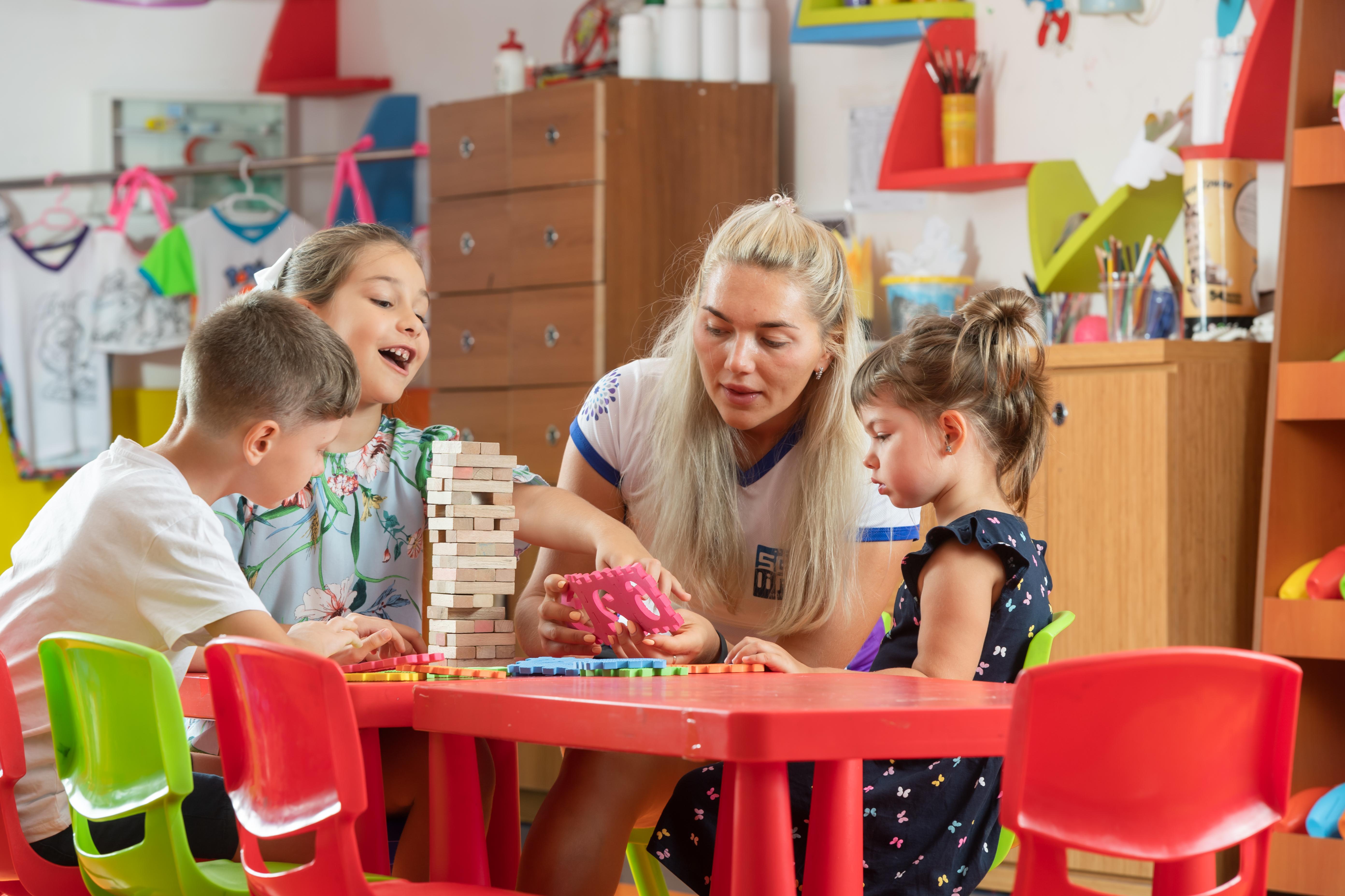 אנטליה Sealife Family Resort Hotel מראה חיצוני תמונה The photo depicts a playful and engaging scene in a classroom or activity space. There are four children sitting at a bright red table, focused on a game. Two girls, one wearing a colorful floral shirt and the other in a dark dress with ruffles, are 