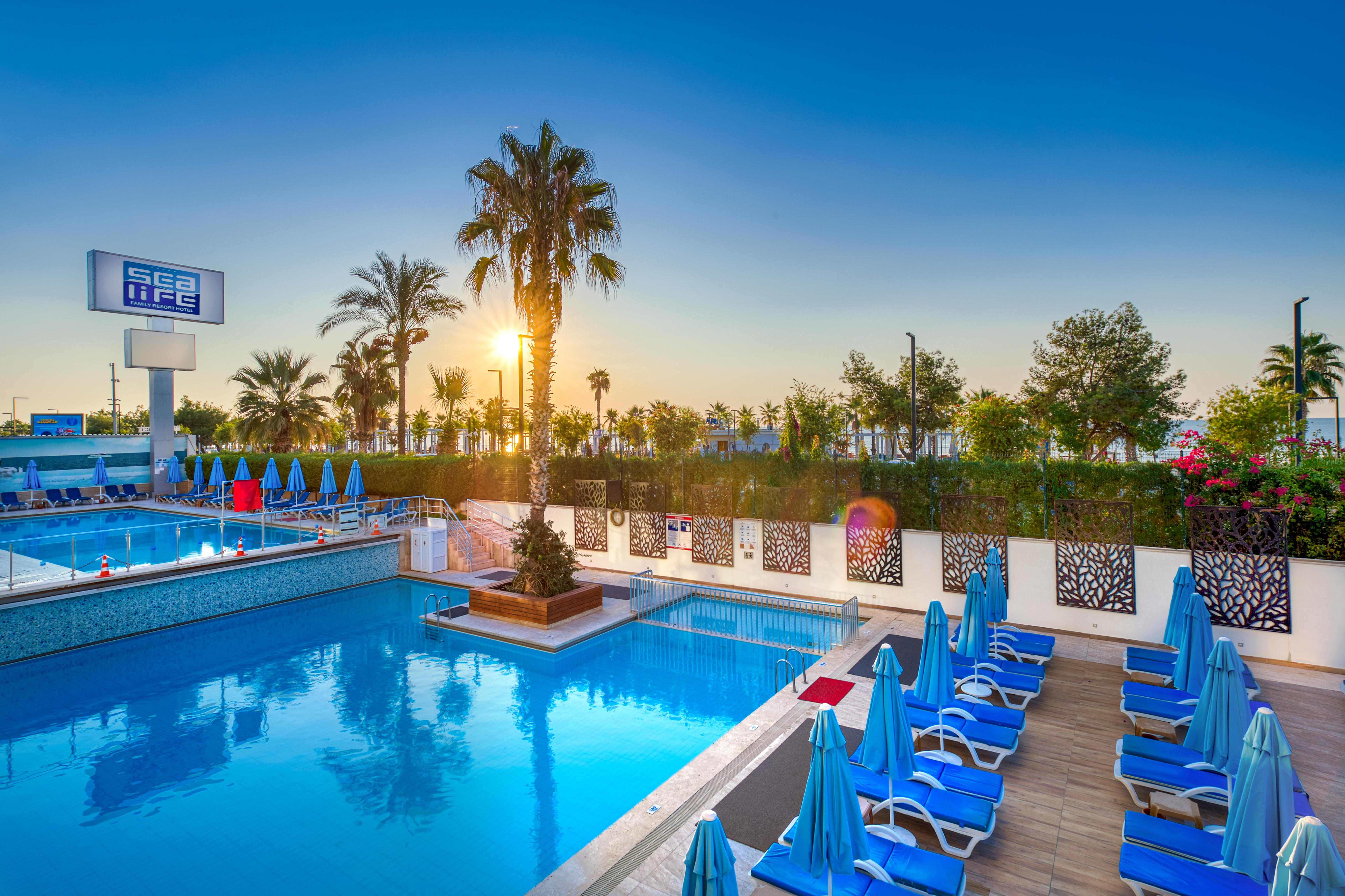 אנטליה Sealife Family Resort Hotel מראה חיצוני תמונה The photo depicts a serene outdoor swimming pool area, illuminated by the warm light of a setting sun. There are several blue lounge chairs arranged around the pool, with palm trees and lush greenery in the background. The sky is clear, showcasing a 