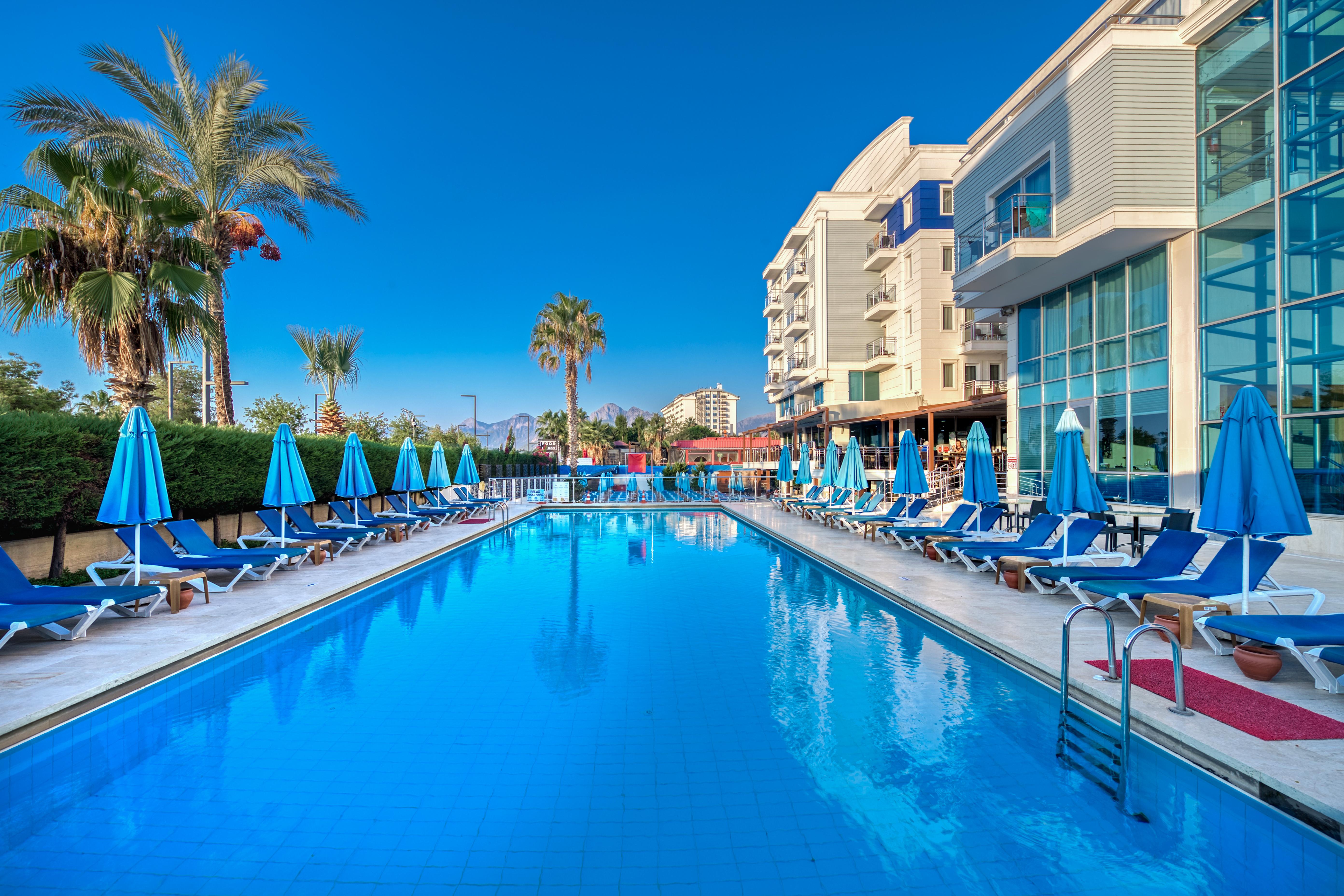 אנטליה Sealife Family Resort Hotel מראה חיצוני תמונה The photo depicts a serene poolside scene at a hotel or resort. In the foreground, a large blue swimming pool reflects the clear sky, with comfortable lounge chairs arranged neatly around it under blue umbrellas. Lush palm trees add to the tropical a