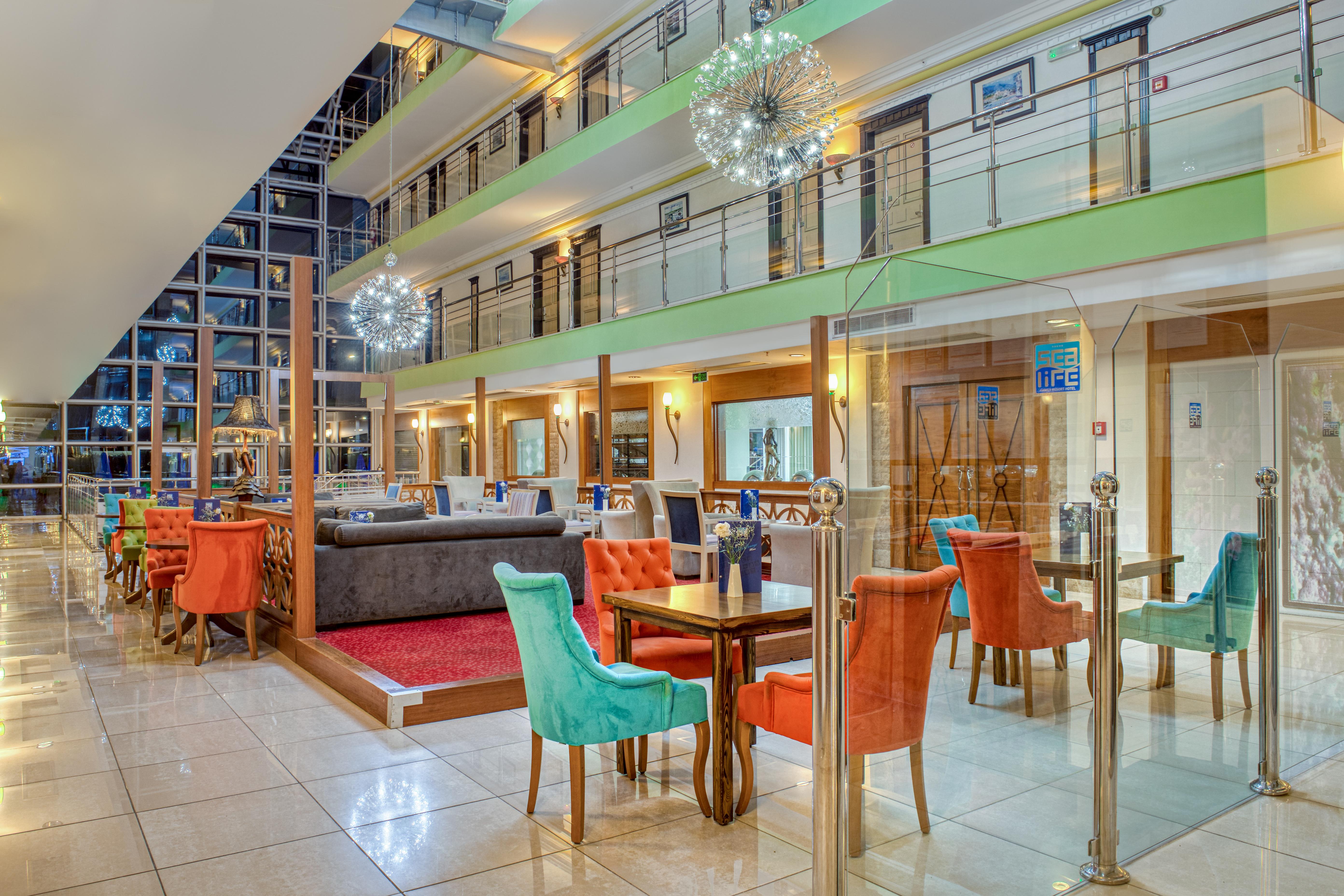אנטליה Sealife Family Resort Hotel מראה חיצוני תמונה The photo shows the interior of a modern hotel lobby or dining area. It features a spacious room with multiple levels visible. The decor includes colorful chairs in orange and turquoise, along with wooden tables. 

There are glass barriers separating
