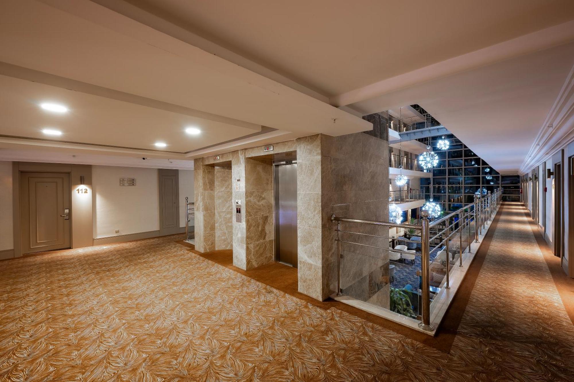 אנטליה Sealife Family Resort Hotel מראה חיצוני תמונה The photo shows a modern hotel corridor. On the left, there are elevators next to a light-colored wall. The floor is covered with a patterned carpet, and there are several ceiling lights illuminating the area. To the right, there is a railing leading