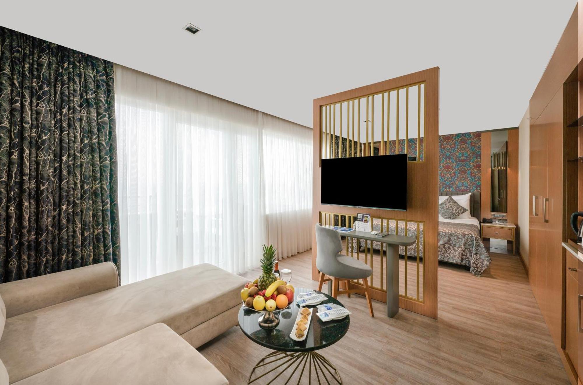 אנטליה Sealife Family Resort Hotel חדר תמונה The photo shows a modern hotel room that features a stylish and functional design. On one side, there is a comfortable light-colored sofa positioned next to a round coffee table adorned with a variety of fruits and snacks. The coffee table has a slee